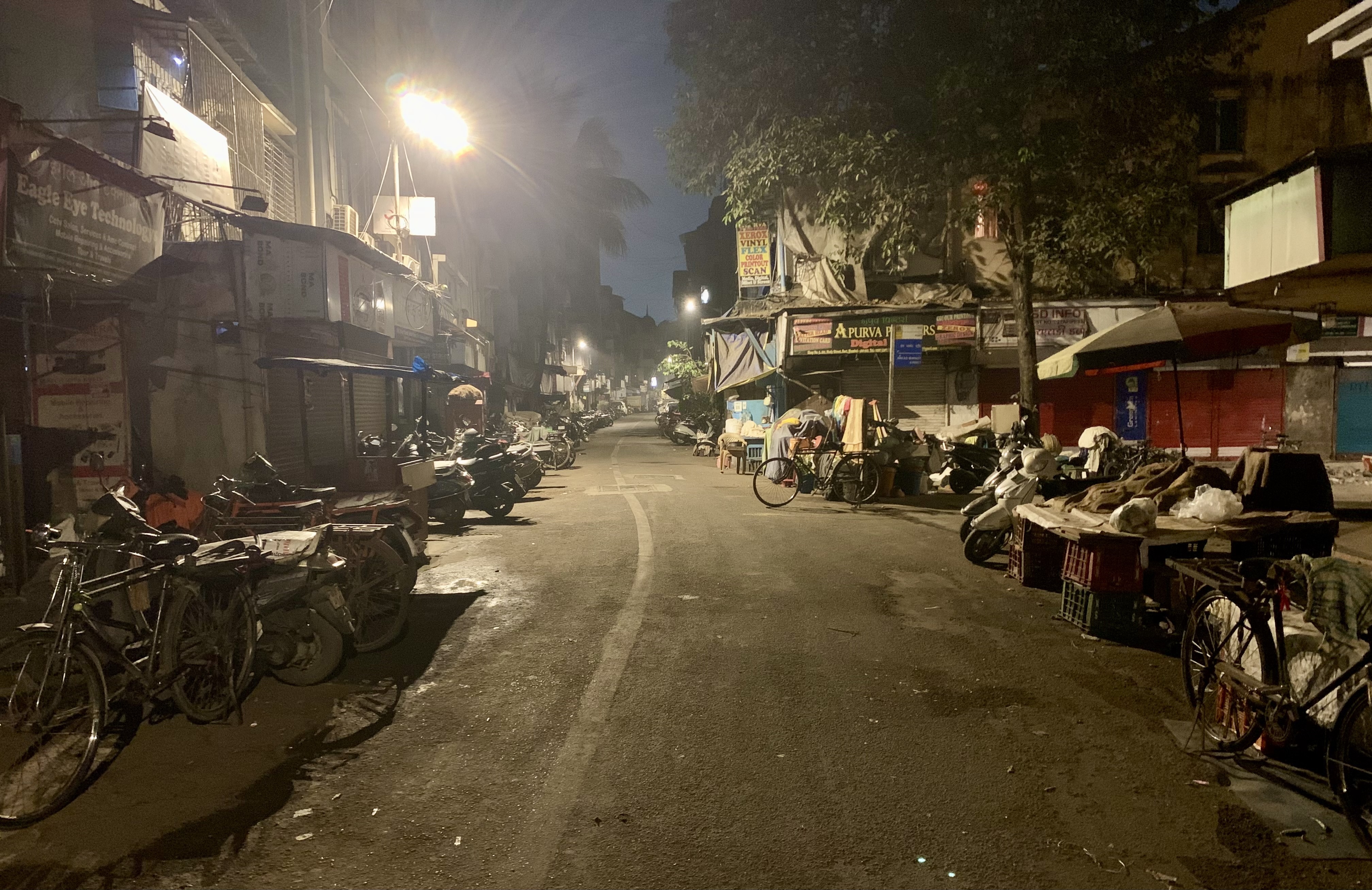 Calle comercial de Mumbai de noche con motos aparcadas, persianas y puestos bajo farolas